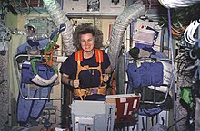 A woman running on a treadmill, anchored by orange straps. The wall behind her has a variety of items including clothing, ventilation hoses and instrument panels affixed to it.