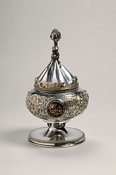 Silver gilt vessel with conical lid. It was intended for use as a ciborium. The vessel is decorated with foliate scrolls and medallions. The knob on the vessel's lid is in the shape of a bunch of grapes.