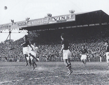 Scène d'un match de football photographiée en noir et blanc. Des joueurs se tiennent prêts à la réception du ballon