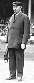 A full-length photograph of a man who looks toward the left side of the picture. His facial expression appears though he may laugh. He is wearing a suit underneath a dark peacoat, as well as a Gatsby-style cap. He holds a protective metal and leather mask in his right hand.