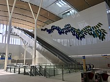 Large shiny white wall with metal escalator casing travelling up alongside it and large blue-green petal-shaped objects attached to the wall