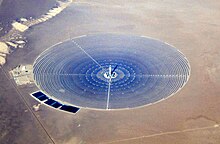 An areal view of a large circle of thousands of bluish mirrors in a tan desert