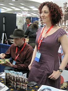 A woman with curly red hair standing behind a desk greetin people