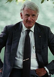 waist high portrait outdoors of smiling white haired man in his sixties hands resting on hips wearing dark suit and tie, and white shirt