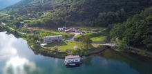 Aerial shot of a lakeside property showing one house built over a lake, a second house behind that with a large yard in front of it, and a third house behind the second, located on a slight incline of land, surrounded by mountains