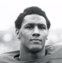 Tight headshot of a dark skinned man with a slightly long afro lookin into the camera. His football shoulder pads are slightly visible.