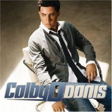 The cover features a man sitting on a white chair wearing an unbuttoned grey shirt, white dress shirt, black tie and blue jeans. The artist's name is placed below him, "Colby O" colored in blue and "Donis" colored in white.