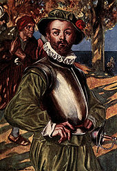 A bearded man, wearing green shirt and doublons, stands with his hands on his hips. The sea is visible in the background. A cuirass (metal armor) protects his upper torso. He is also wearing a green hat with a red rose and a white ruff on his neck. A rapier hangs ready at his left hip. Behind him on his right, a man is walking by, carrying a laden rucksack over his shoulder.