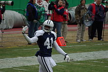 An American football player leans forward. He is not wearing pads but is wearing his white jersey over a long-sleeved gray shirt.