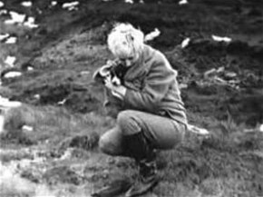 A crouched blonde woman in thick jacket, trousers, and boots, holding a small dog.