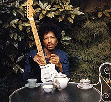 A color image of a man sitting at a table in the outdoors holding a black guitar.