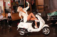 Picture of a white Vespa scooter standing in a street corner. A blonde woman is sitting on the scooter. She is wearing a white short dress and white-heeled shoes. Her bare right leg is put up on the handle of the Vespa. Behind her, on the left, the bonnet of a indigo colored car is visible, while the bowed down head of a man wearing a white hat is visible on her right