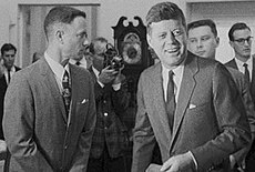Black-and-white film screenshot showing the main character on the left looking towards another man, President Kennedy, on the right. Kennedy is smiling and looking to his left. In the background several men are looking in different directions and one is aiming a camera.