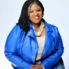 An image of a woman sitting on a chair, smiling for a photo against a white background while wearing a bright blue blazer.