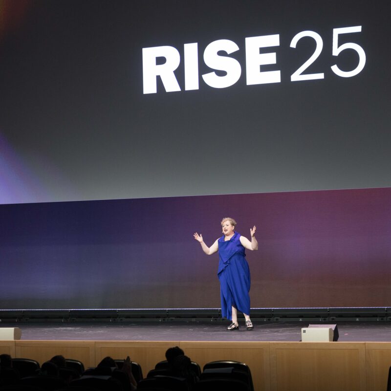 A speaker in a vibrant blue dress stands on a large stage, gesturing expressively with her arms raised. The stage backdrop is a gradient of dark colors, and the word "RISE25" is prominently displayed on the screen behind her. The audience is visible in the foreground, seated and watching the presentation.