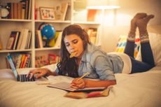 Teenage girl lying on bed in her room and studying. She is using computer to help her for researching. Eating cookie for snack. Evening or night with beautiful yellow lights lightning the scenes.