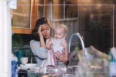 A mum looking stressed while holding a baby, pictured through a kitchen window