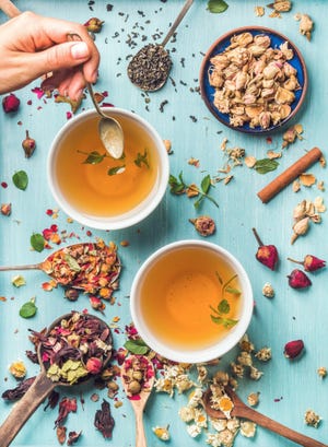 two cups of healthy herbal tea with mint, cinnamon, dried rose, camomile flowers in spoons and man's hand holding spoon