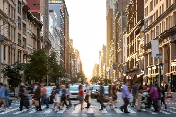 Diverse crowd of people walking on a sunny day across a busy intersection of a large city.