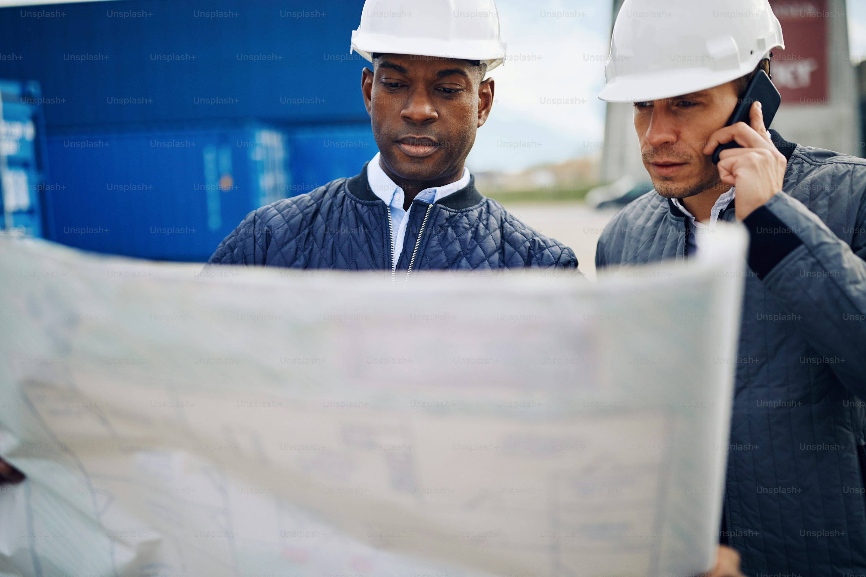 Two engineers standing by freight containers on a large commercial shipping dock reading a blueprint and talking on a cellphone