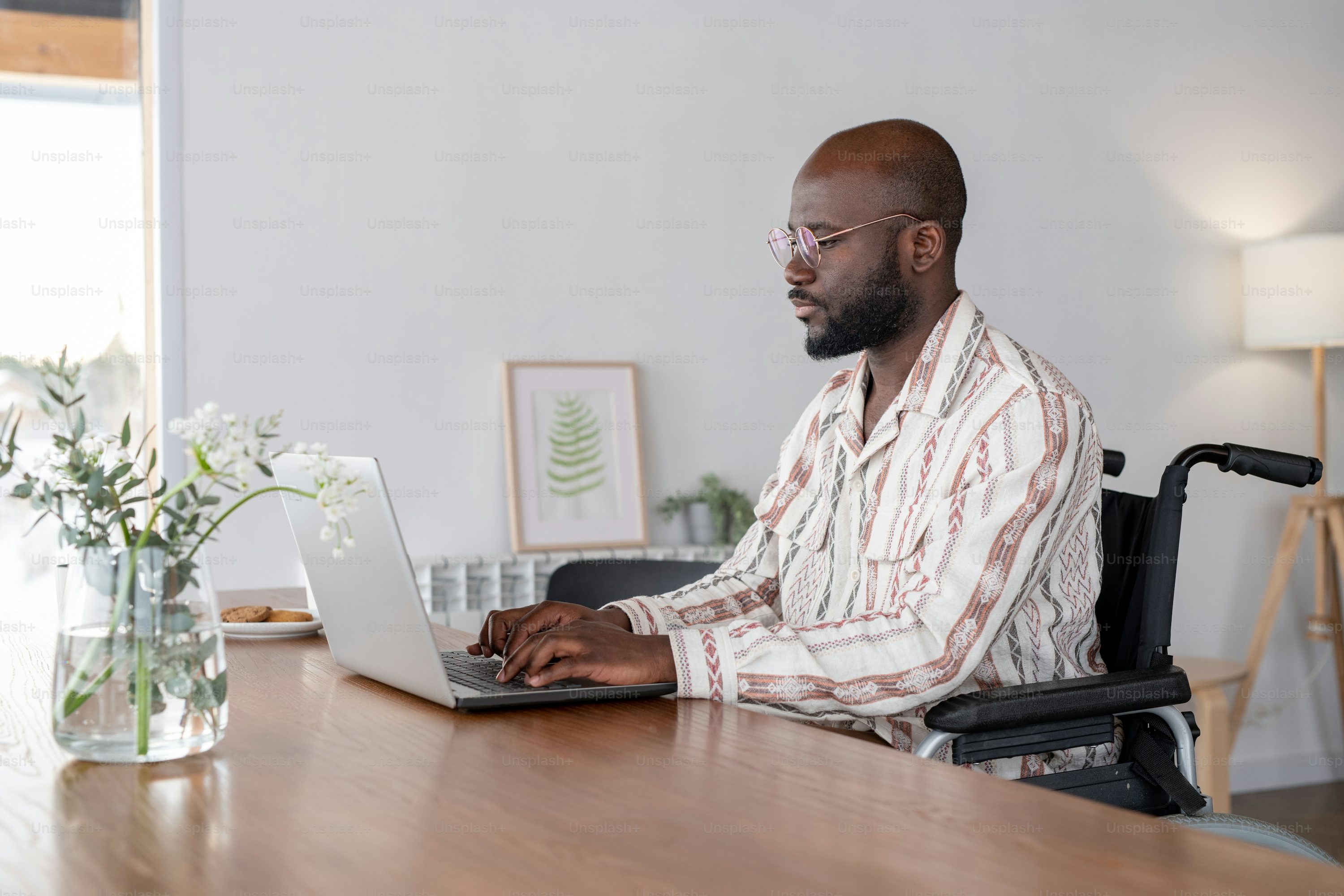 Young serious businessman or freelancer in wheelchair sitting by table in living room and looking at laptop screen while typing