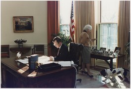 President Bush works at his desk in the Oval Office as Mrs. Bush looks at photographs on the table behind the Oval... - NARA - 186389.tif