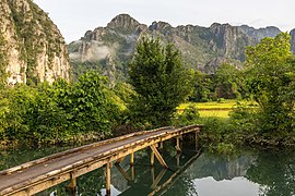 Three-quarter view of a wooden footbridge over a lagoon, trees and mountains in Vang Vieng, Laos.jpg