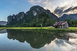 Water reflection of karst mountains, wooden hut, trees and colorful clouds at sunset with green paddy fields, Vang Vieng, Laos.jpg