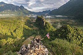 Man on a motorcycle holding a flag of Laos in front of green karst peaks at the top of Mount Nam Xay, Vang Vieng, Laos.jpg