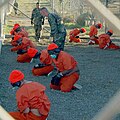 Detainees in orange jumpsuits sit in a holding area under the watchful eyes of Military Police at Camp X-Ray at Naval Base Guantanamo Bay, Cuba, during in-processing to the temporary detention facility on Jan. 11, 2002. The detainees will be given a basic physical exam by a doctor, to include a chest x-ray and blood samples drawn to assess their health. DoD photo by Petty Officer 1st class Shane T. McCoy, U.S. Navy.