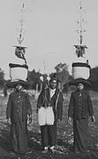 COLLECTIE TROPENMUSEUM Een man met een versierde draagstok met manden wordt geflankeerd door twee Batak vrouwen die ritueel versierde zakken op hun hoofd dragen TMnr 60043414.jpg