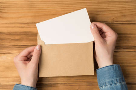 First person top view photo of female hands holding open craft paper envelope with white card on isolated wooden desk background with blank space
