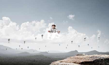 Young little boy keeping eyes closed and looking concentrated while meditating on cloud in the air with beautiful and breathtaking landscape on background