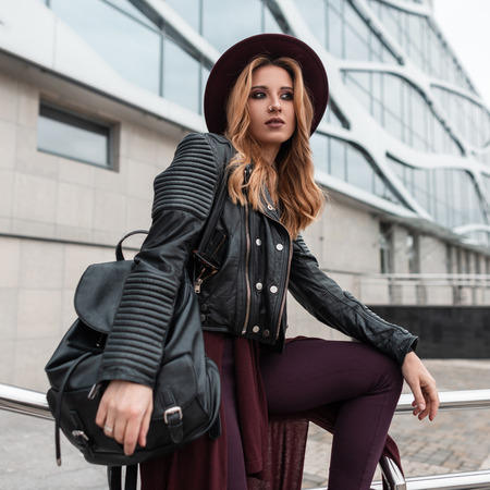 Young pretty attractive woman in black leather jacket in pants with a stylish backpack in an elegant hat posing sitting near a modern building in the city fashionable red haired girl enjoys vacation