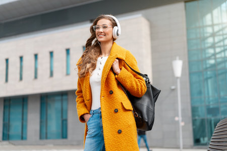 Woman wear eyeglasses listen music headphone walking outdoor city street dressed stylish yellow coat and black backpack smile caucasian female 30s enjoy podcast or audio books outside