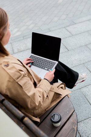 A young unrecognizable woman is working on a laptop sitting on a bench near the office freelancer while working black screen on laptop mockup Stock Photo