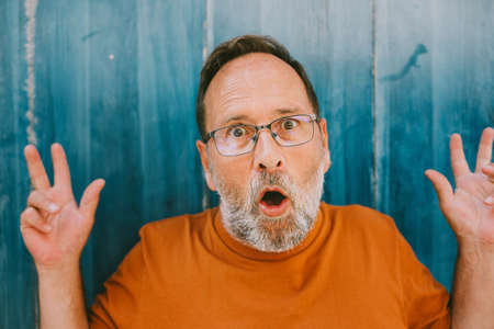 Retrato al aire libre de un hombre de edad muidle sorprendido, con camiseta naranja y anteojos, posando sobre fondo azul