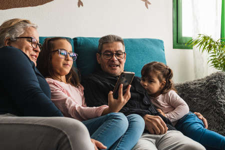 Abuelo y abuela, sentados en el sofá de casa con sus nietas, viendo un video en el teléfono móvil, mientras se divierten juntos.