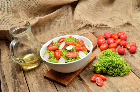 Green salad lollo biondo with tomatoes and radishes in a white bowl on wooden table a pitcher of oil in the background Stock Photo