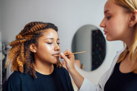 Caucasian blonde female master makes makeup for her dark skinned client in a beauty salon a pretty african american blue eyes young woman is putting lipstick on her lips concept beauty and spa