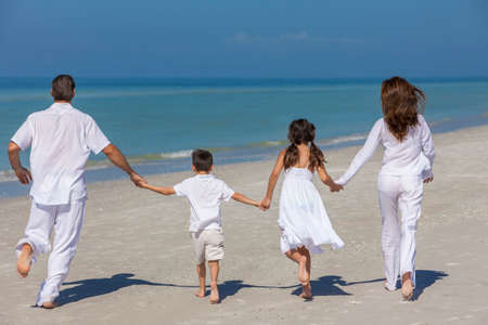 Vista trasera de la familia feliz de madre, padre y dos hijos, hijo e hija, corriendo la mano y divertirse en la arena de una playa soleada