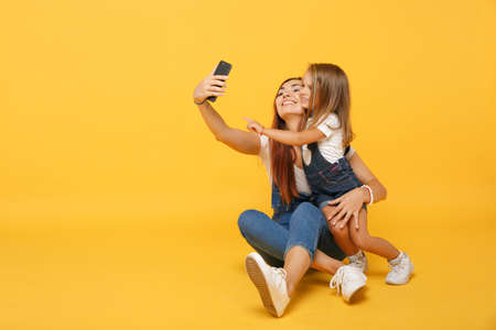 Mujer con ropa ligera haciendo selfie con una linda niña de 4 a 5 años. Mamá pequeña hija aislada en un retrato de estudio de fondo amarillo. Concepto de infancia de paternidad familiar del Día de la Madre. Foto de archivo