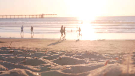 Gente caminando en la playa de arena del océano por el muelle al atardecer, costa de California, Estados Unidos. Siluetas familiares desenfocadas irreconocibles a la luz del sol brillante, vacaciones en la costa bajo el sol. Ondas de agua de mar iluminadas por el sol.