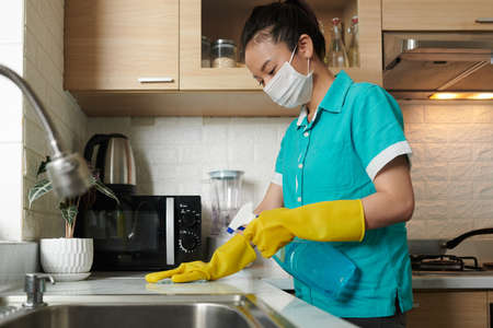 Trabajador de servicio de limpieza profesional en uniforme con máscara médica al limpiar la cocina con detergentes tóxicos