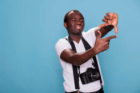 Młody facet z urządzeniem dslr gestykuluje robiąc zdjęcie rękami pewny siebie profesjonalny fotograf robi gest ramki rękami, mając nowoczesny aparat na niebieskim tle ujęcie studyjne