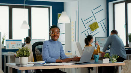 Retrato de una mujer afroamericana sonriente que trabaja en el margen de ganancias y las estadísticas de venta usando una computadora portátil sentada en el escritorio en una oficina de inicio ocupada. empleado de negocios casual feliz con el resultado del trabajo.