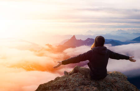 El hombre aventurero con las manos abiertas está disfrutando del momento en la cima de una montaña. Compuesto de fantasía. Cielo del atardecer o del amanecer. Paisaje de la Columbia Británica, Canadá.