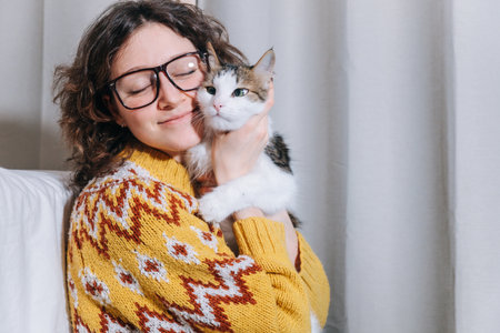 Linda chica independiente con gafas y un suéter amarillo está trabajando en casa en una computadora portátil sentada en el suelo y abrazando a su gato blanco