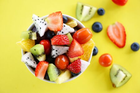 Fruit salad bowl fresh summer fruits and vegetables healthy organic food strawberries orange kiwi blueberries dragon fruit tropical grape pineapple tomato lemon on yellow background