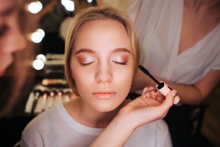 Close up beautiful young blonde woman face she has eyeshadows on eyeline make up artist use mascara hairdresser work behind